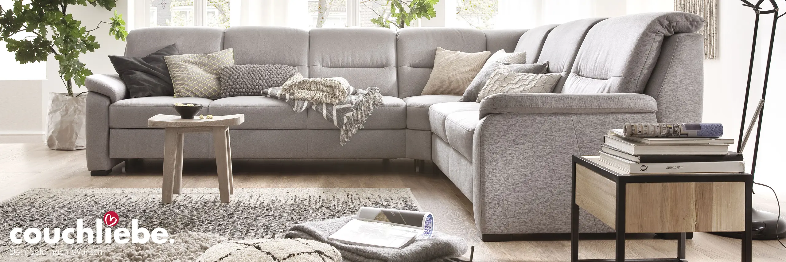 Graues Ecksofa mit Kissen und Decke, runder Holz-Couchtisch auf Teppich, Beistelltisch mit Büchern rechts und Pflanze links im Wohnzimmer, Text couchliebe